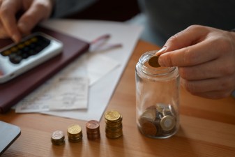 Imagem de uma mão colocando moedas dentro de um pote ao lado de várias pilhas de moeda e uma calculadora. Imagem para a matéria sobre viver de renda com 500 mil