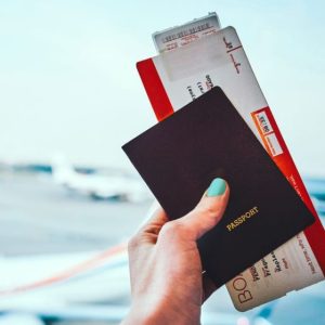 Imagem para a matéria sobre como comprar passagens aéreas baratas em que aparece uma mão segurando um bilhete de avião Ao fundo a pista de um aeroporto com um avião.