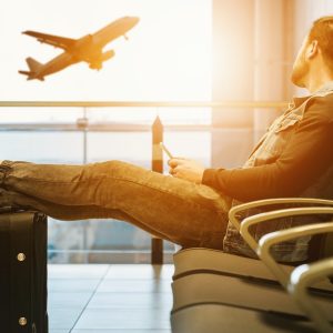 Imagem sobre destinos brasileiros baratos em que aparece um homem sentado em um aeroporto com as pernas esticadas em cima da mala olhando um avião subir.