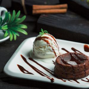 Foto de um prato branco, com um bolinho de chocolate e ao lado uma bola de sorvete. No canto da foto há uma xicara com uma planta.