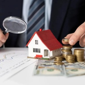 Foto de uma pessoa segurando uma lupa enquanto empilha moedas do lado de uma maquete de uma casa. A matéria trata dos fundos imobiliários baratos.