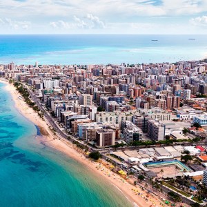 Foto de uma paisagem vista de cima de Maceió, em Alagoas. De um lado há um mar bem azulado. Do outro, uma cidade com prédios e casas.