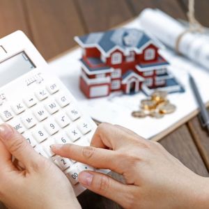 Imagem para a matéria sobre como investir em imóvel em que aparecem duas mãos segurando uma calculadora e ao fundo uma maquete de uma casa com uma pilha de moedas na frente.