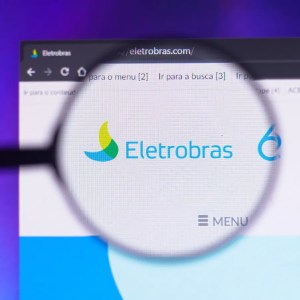 Foto de uma lupa sobre o logo da Eletrobras em seu site oficial. A matéria mostra quanto renderam 100 ações ELET3 e ELET6.