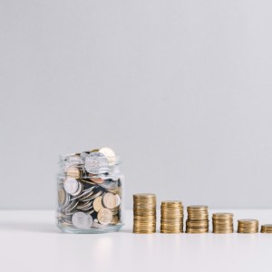 Imagem traz um pote de vidro com moedas, seguido de pilhas de moedas, para ilustrar reportagem sobre as desvantagens do CDB