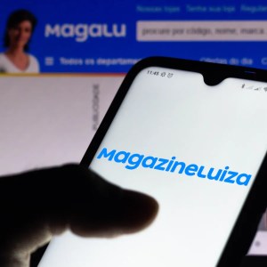 Foto de uma mão segurando um celular com a imagem do logo do Magazine Luiza. A matéria calcula quando renderam as ações (MGLU3) no curto e médio prazo