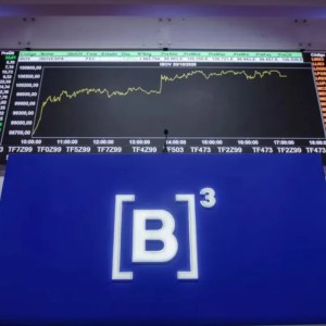 Foto de um painel da bolsa de valores brasileira, a B3 (B3SA3). Em um telão com fundo azul, há um escrito "B3". Ao fundo, um painel com números de ações.