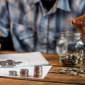 Imagem para a matéria sobre investimento a longo prazo em que apraece um homem colocando moedas em um pote. Em cima da mesa também estão papeis e pilhas de moedas.