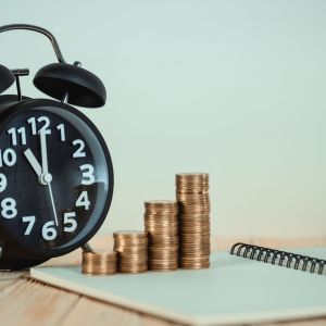 Imagem para a matéria sobre investir em renda fixa a curto prazo em que aparece um relógio de ponteiro em cima de uma mesa. Ao lado uma pilha crescente de moedas e um caderno aberto.