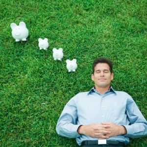 Foto de um homem com uma camisa azul deitado em um gramado com os olhos fechados. Ao lado dele, em formato de pensamento, estão quatro cofrinhos brancos.
