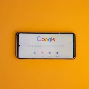 Foto de celular na horizontal com tela inicial do Google. Celular está sobre um fundo totalmente laranja.