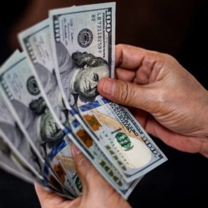 Foto de uma mão segurando notas de dólar americano. A matéria explica a alta do dólar em abril de 2024