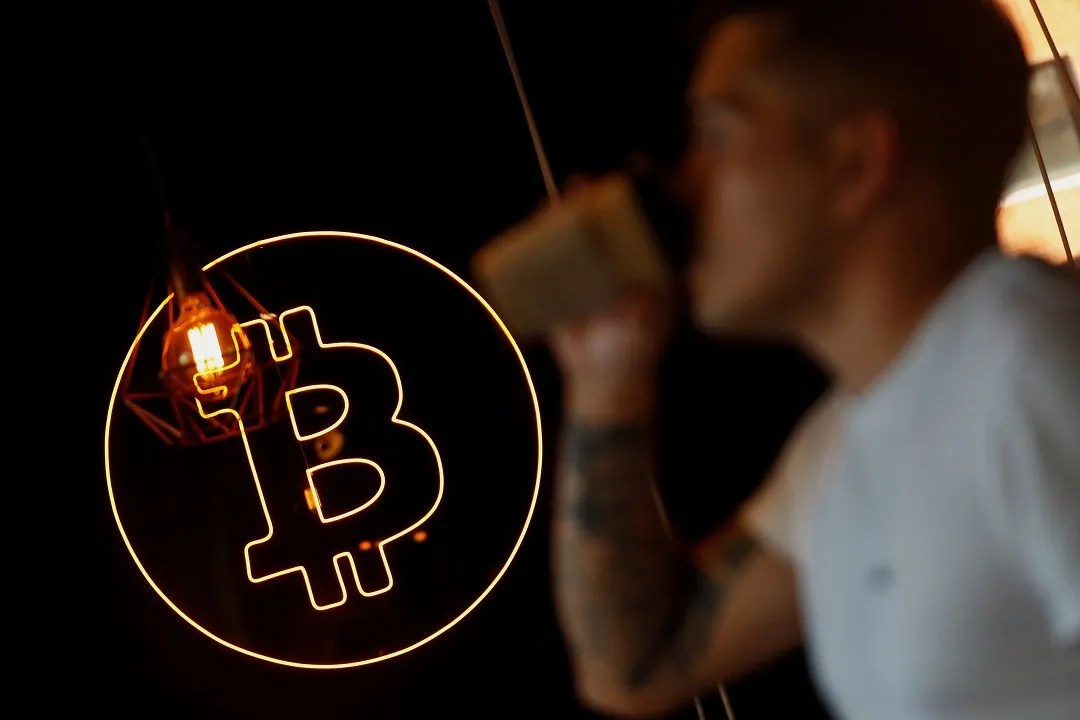 Foto do símbolo do bitcoin, com uma moeda laranja e uma letra B dentro. Ao fundo, há o perfil desfocado de um homem tomando um café.