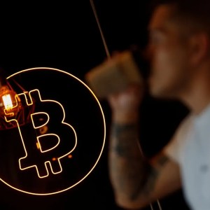 Foto do símbolo do bitcoin, com uma moeda laranja e uma letra B dentro. Ao fundo, há o perfil desfocado de um homem tomando um café.