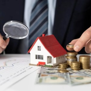 Foto de uma pessoa com uma lupa na mão esquerda e moedas na mão esquerda, com uma casa no meio. A matéria fala sobre investir em fundos imobiliários.