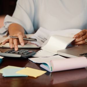 Imagem para a matéria sobre quanto rende R$ 1 milhão na conta corrente em que aparece uma mulher fazendo cálculos em uma calculadora e vários papeis em volta.