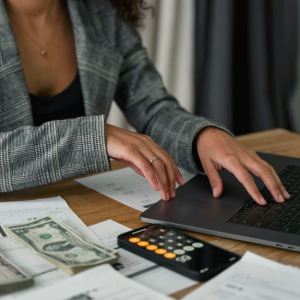 Foto de uma mulher com um casaco cinza, mexendo em um computador. Ao lado dela há uma calculadora e algumas notas de dinheiro.