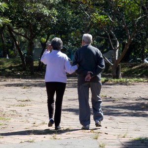 Imagem em área aberta, local arborizado, mostra casal de idosos caminhando. Ambos são fotografados de costas e usam agasalhos. Tema da matéria é 