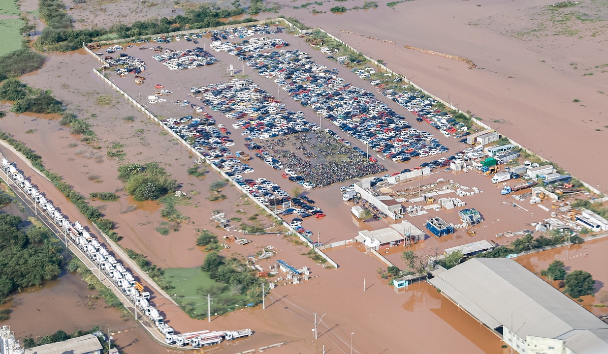 Foto de uma área afetada pelas enchentes no Rio Grande do Sul, mostra casas e instalações submersas. A matéria descreve o impacto do evento no PIB do Brasil.