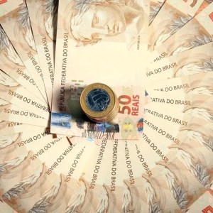 Imagem para matéria sobre investimentos para viver de renda em que aparecem várias notas de 50 reais formando um círculo. Em cima de uma das notas está uma pilha de moedas de 1 real.