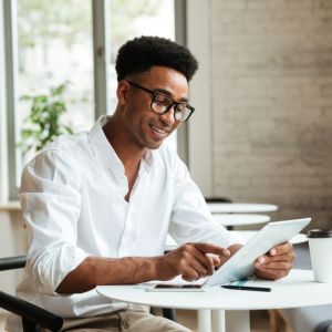 Imagem para matéria sobre quanto rende 100% do cdi em que aparece um homem negro de óculos e roupa branca sentado em uma mesa olhando um tablet.
