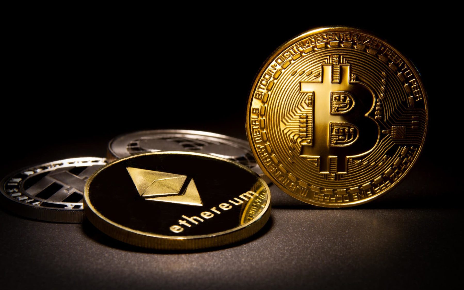 Foto de uma moeda de bitcoin, ou BTC, dourada ao lado de uma moeda de Ethereum, ou ETH, também dourada, sobre um fundo preto. A matéria explica se vale a pena investir em um ou outro.