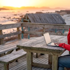 Nômade digital sentado ao ar livre na mesa na praia com um laptop sozinho