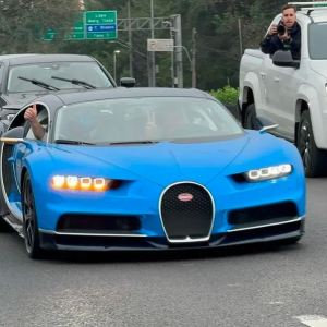 Foto de um caro azul de modelo esportivo, andando em uma rua. Ao lado, há outro carro com fotógrafos.