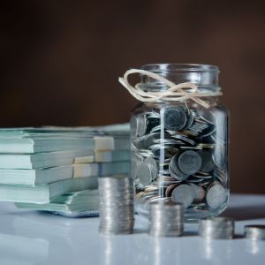 Foto de um pote de vidro transparente cheio de moedas. Ao lado, há um bolo de notas de dinheiro.