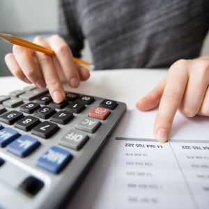 Imagem para matéria sobre quanto rendem R$ 300 mil no CDB por mês em que aparece uma mão de uma mulher branca fazendo cálculos em uma calculadora. Ao lado, um papel com tabelas e números.