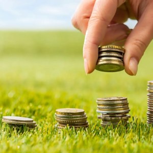 Imagem para matéria sobre como investir no agronegócio em que aparecem várias pilhas de moedas e uma mão empilhando mais moedas. Essas moedas estão em um campo verde.