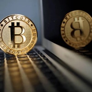 Foto de uma moeda de bitcoin dourada sobre um computador. A matéria mostra quanto renderam R$ 5 mil em bitcoin.