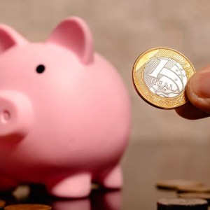 Imagem de um cofrinho em formato de porco na cor rosa. Ao lado, a mão de uma pessoa branca segura uma moeda de 1 real.