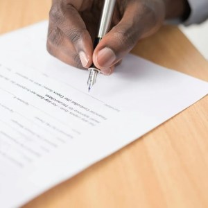 Imagem da mão de um homem negro assinando um documento em cima de uma mesa.