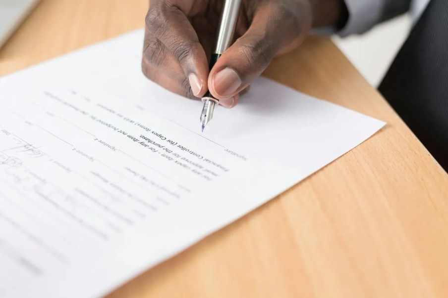 Imagem da mão de um homem negro assinando um documento em cima de uma mesa.